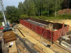 Szczegółowe ujęcie szalunku fundamentowego z czerwonych paneli, przygotowanego do zalewania betonem, widoczna pompa do betonu i zbrojenie, teren budowy w otoczeniu zieleni.