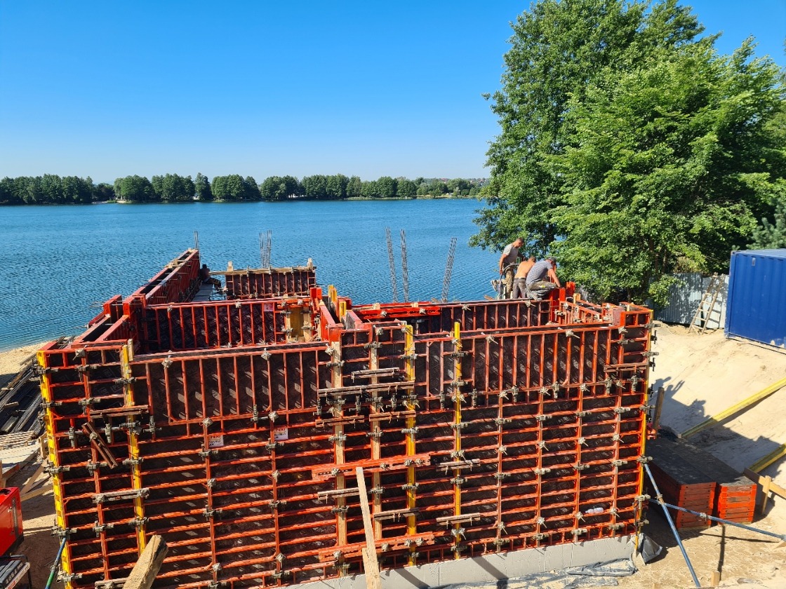 Budowa fundamentów domu przy jeziorze, widoczne szalunki i zbrojenia, pracownicy w trakcie montażu, w tle zbiornik wodny i zieleń.