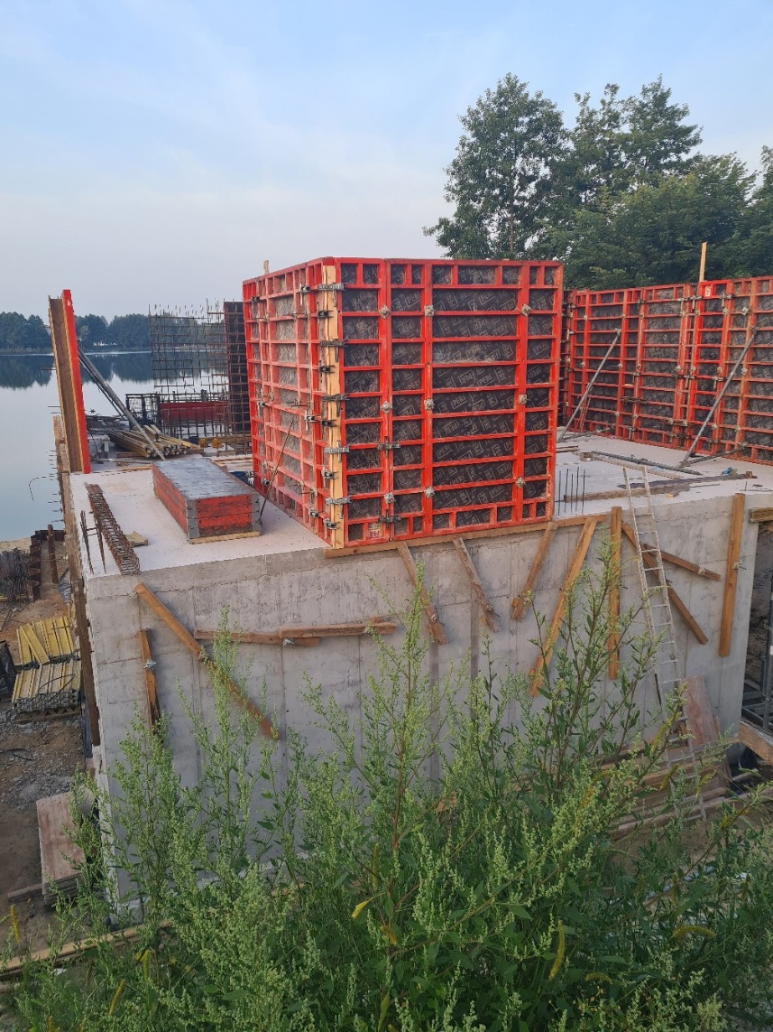 Wznoszenie żelbetowej konstrukcji przy jeziorze, widoczne szalunki w kolorze pomarańczowym oraz zbrojenie, w tle drzewa i woda.