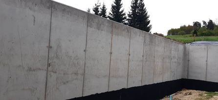 Świeżo wylane, szalowane ściany fundamentowe z betonu, zabezpieczone czarną izolacją przeciwwilgociową, widoczne ślady po szalunkach, w tle drzewa iglaste i trawiasta skarpa.