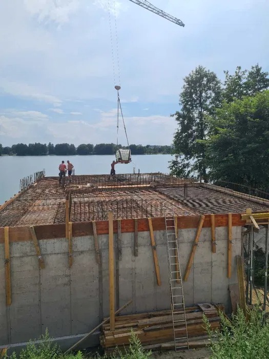 Wznoszenie żelbetowej konstrukcji budynku nad brzegiem jeziora, widoczny dźwig podający materiały, zbrojenie na górze konstrukcji i szalunki na betonowych ścianach.