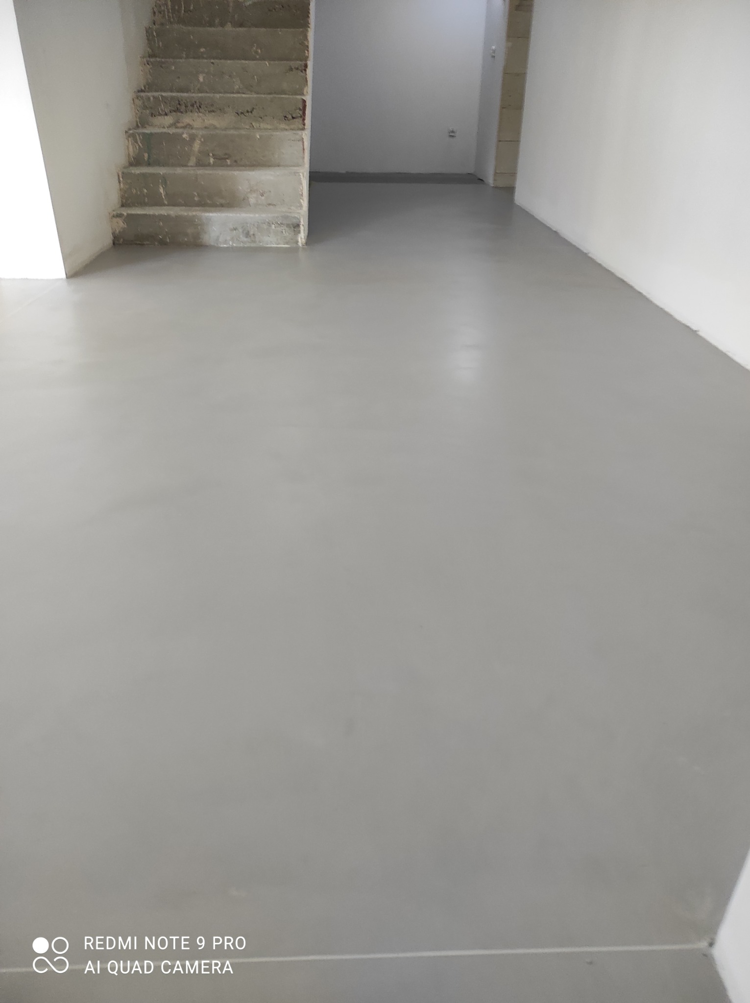 Gładka, szara posadzka w minimalistycznym wnętrzu z surowymi betonowymi schodami i białymi ścianami.