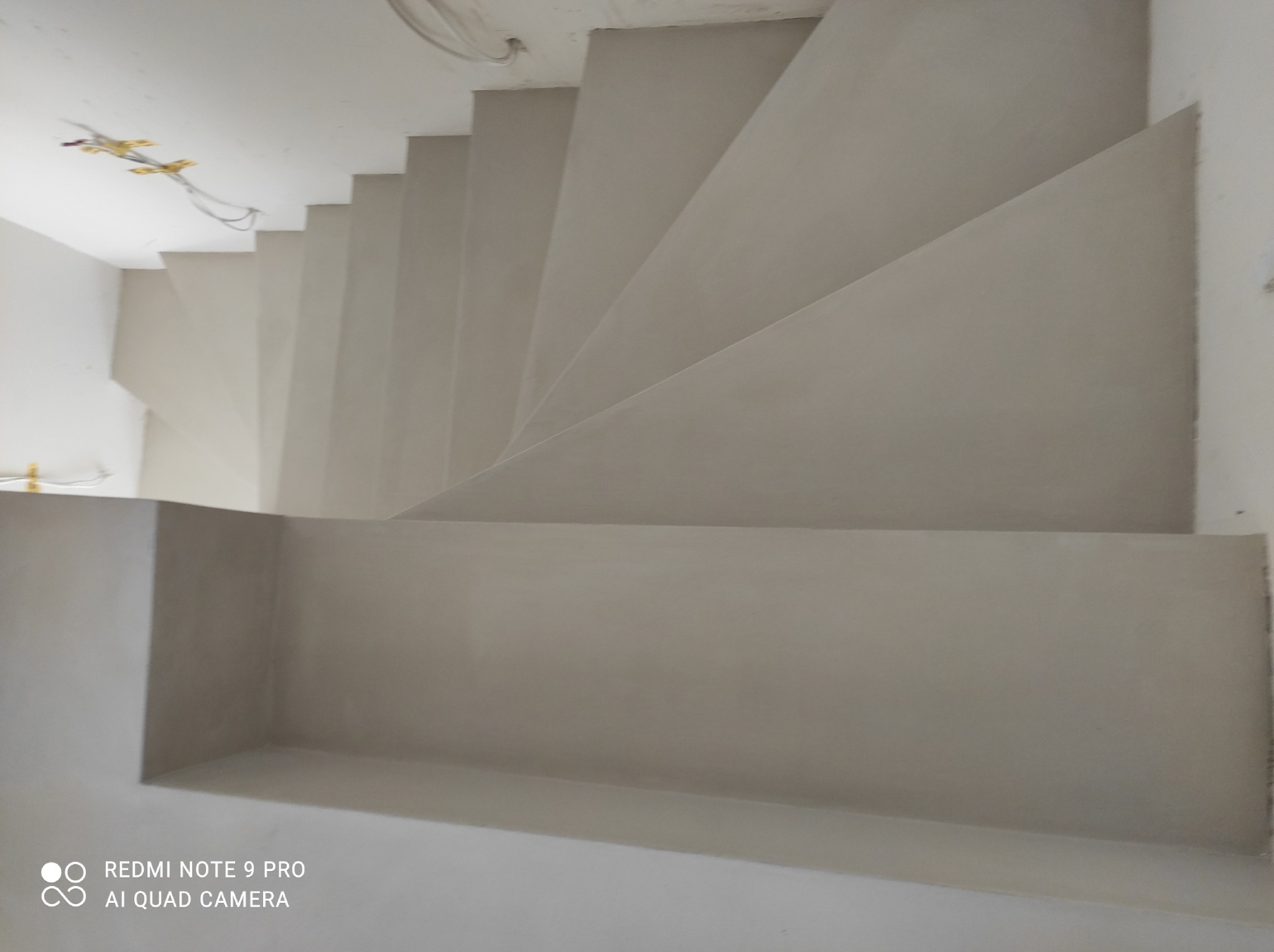 Schody wewnętrzne wykończone jednolitą, jasną powierzchnią, prawdopodobnie mikrocementem, widok z góry, z widocznymi przewodami elektrycznymi na suficie.