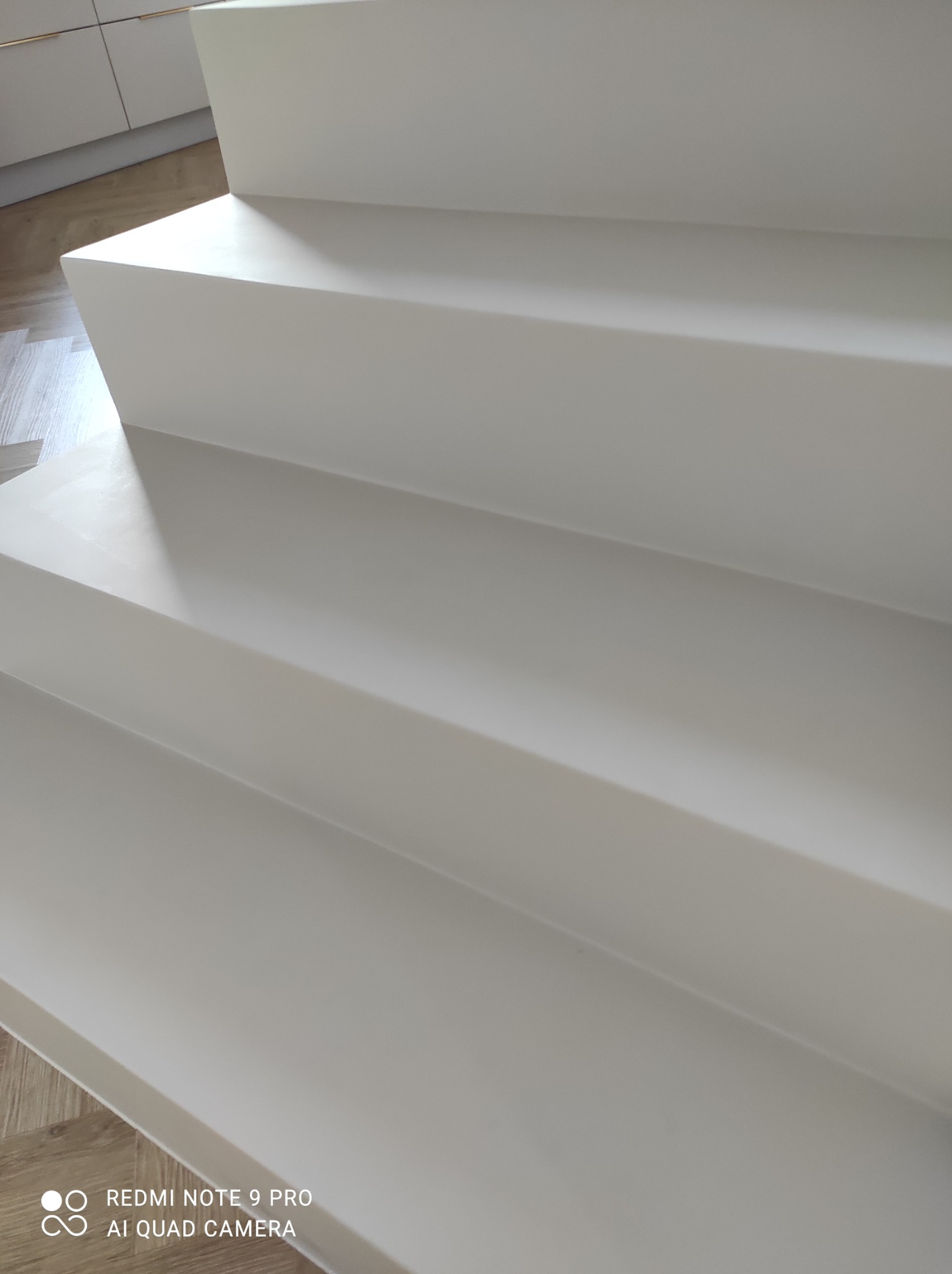 Minimalistyczne, białe schody z gładką powierzchnią, oświetlone naturalnym światłem, z widocznym fragmentem drewnianej podłogi na górze.