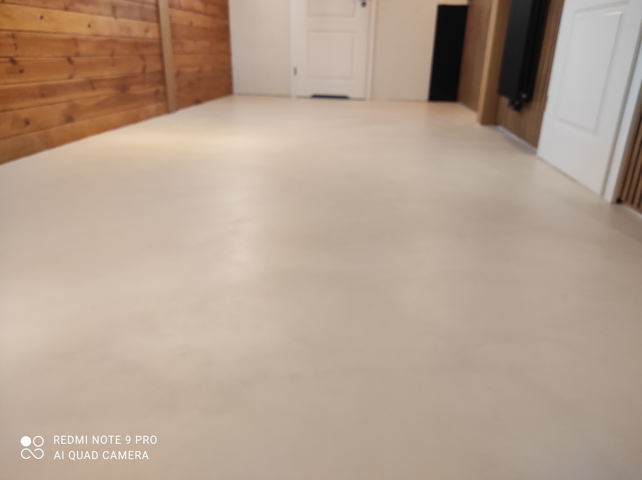 Jasna, jednolita posadzka w odcieniach beżu, wykonana z mikrocementu, widoczna w pomieszczeniu z drewnianą boazerią i białymi drzwiami.