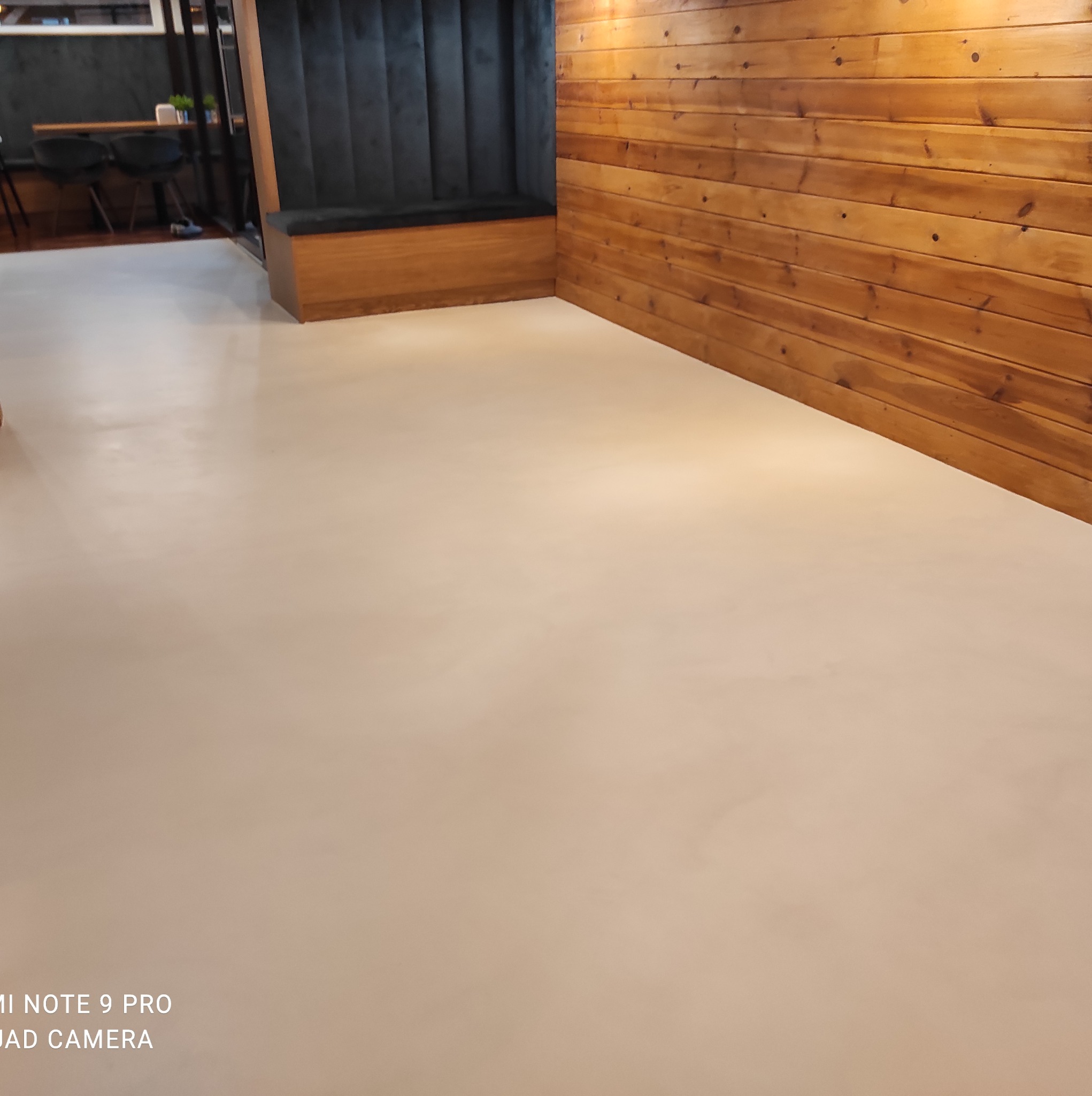 Beżowa, gładka posadzka w lokalu z drewnianą ścianą i czarną, tapicerowaną wnęką do siedzenia.