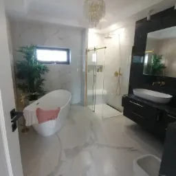 Jasna łazienka z marmurowymi płytkami, wolnostojącą wanną z różowym ręcznikiem, przeszkloną kabiną prysznicową ze złotymi detalami i czarną szafką pod umywalką.