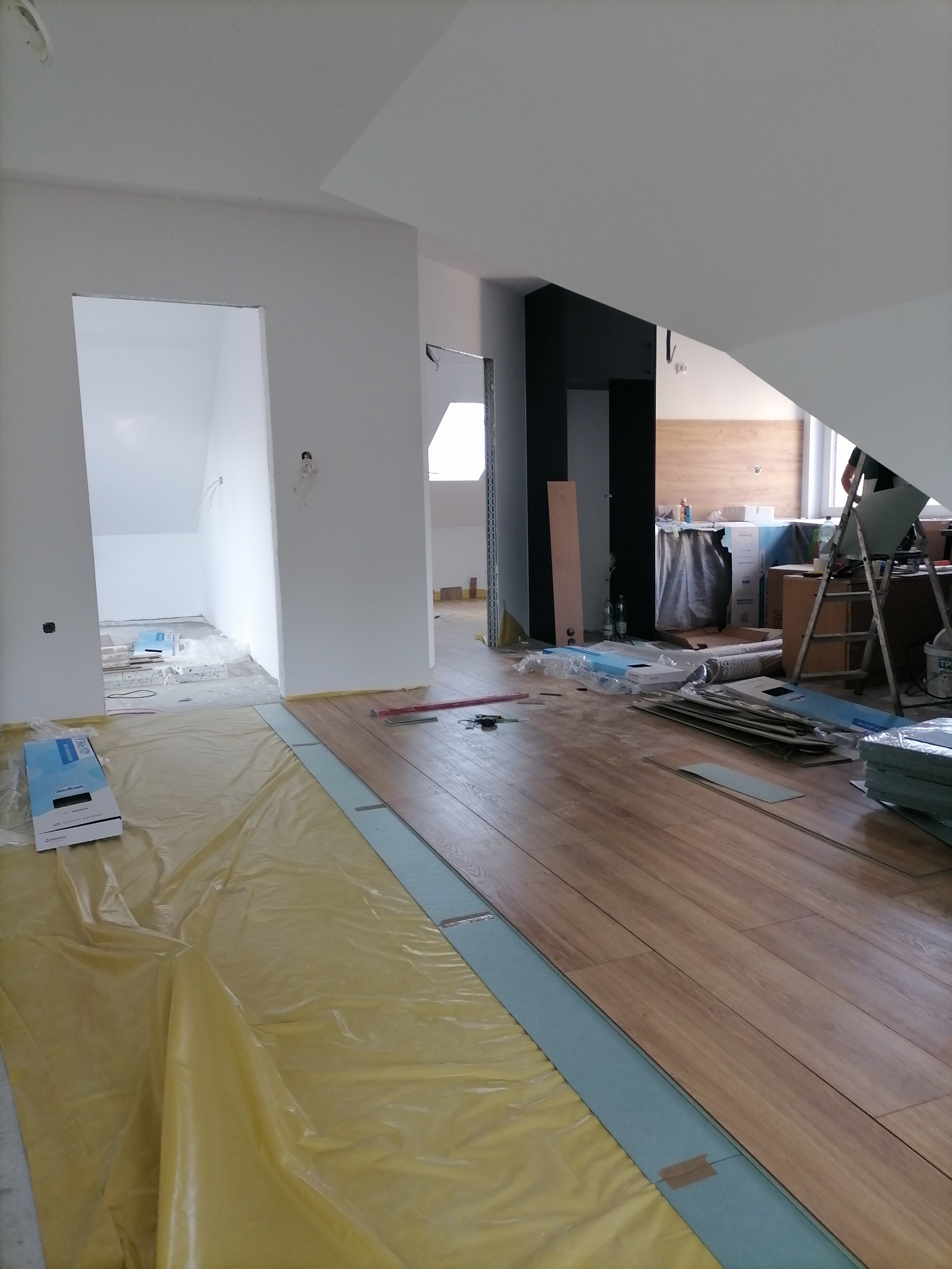 Montaż paneli podłogowych w jasnym wnętrzu z widoczną folią ochronną, fragmentem drabiny i przygotowanymi materiałami.