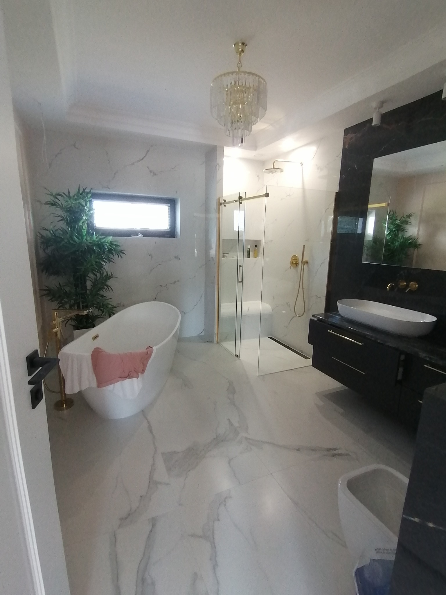 Jasna łazienka z marmurowymi płytkami, wolnostojącą wanną z różowym ręcznikiem, przeszkloną kabiną prysznicową ze złotymi detalami i czarną szafką pod umywalką.