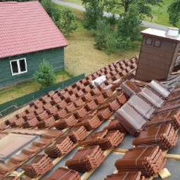 Widok z góry na dach w trakcie układania brązowej dachówki ceramicznej; w tle widoczny zielony budynek z czerwonym dachem i komin.