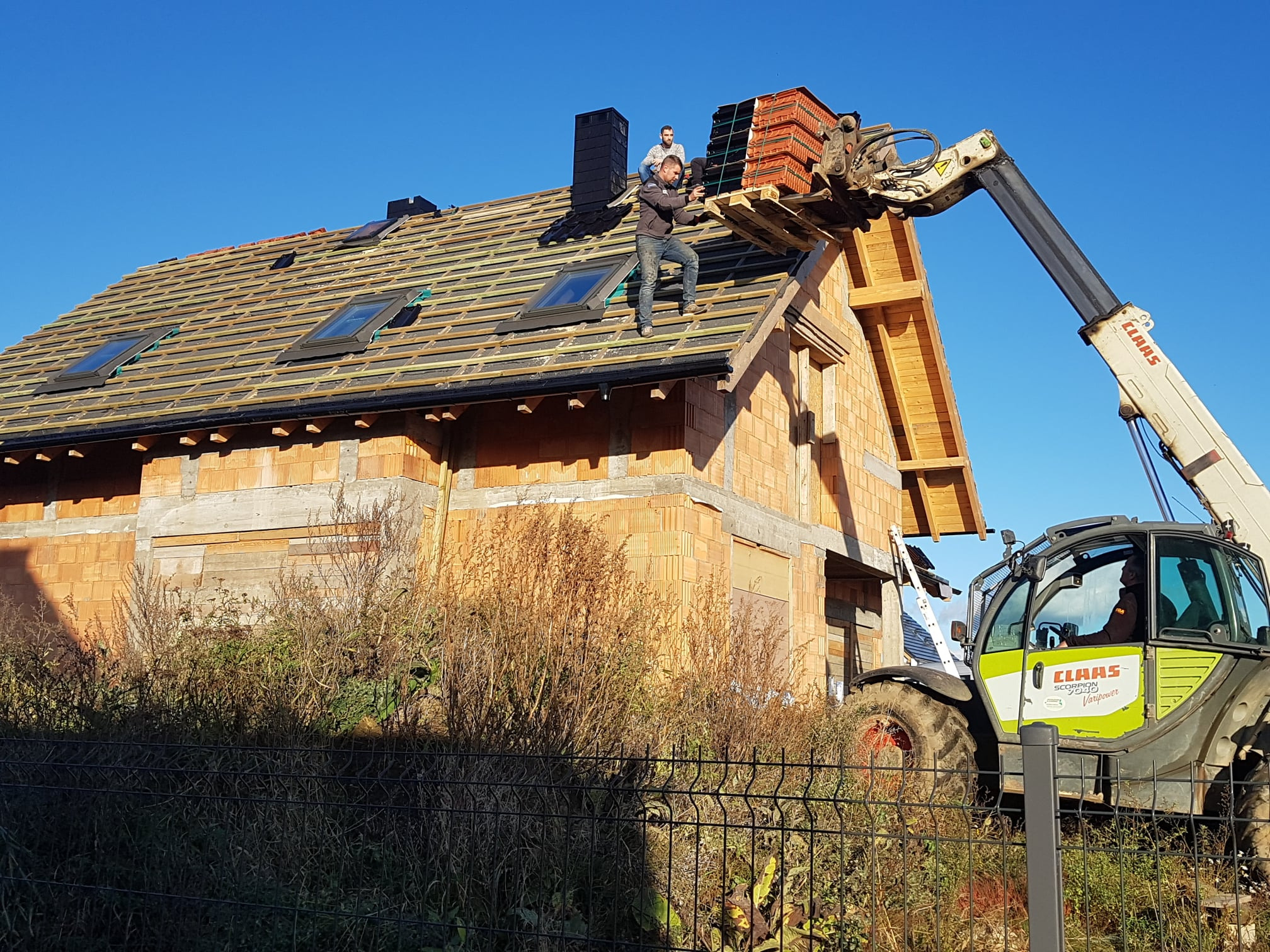 Dwóch dekarzy na dachu domu w trakcie układania dachówki, materiał podawany przez podnośnik teleskopowy Claas, widoczne okna dachowe.