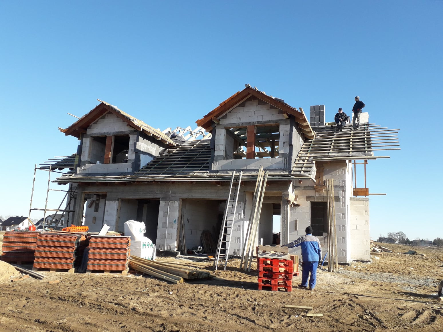 Budowa domu jednorodzinnego w stanie surowym otwartym: murowane ściany, częściowo wykonana konstrukcja dachu z łatami, stosy dachówki na paletach, pracownicy na dachu.