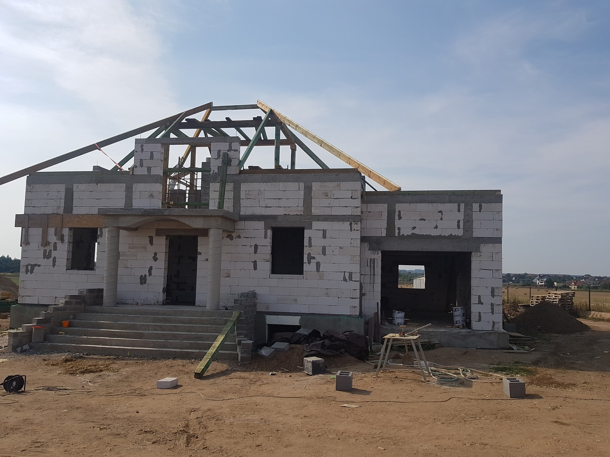 Budowa domu jednorodzinnego w stanie surowym otwartym z widoczną konstrukcją więźby dachowej i schodami wejściowymi, teren budowy z materiałami i narzędziami.