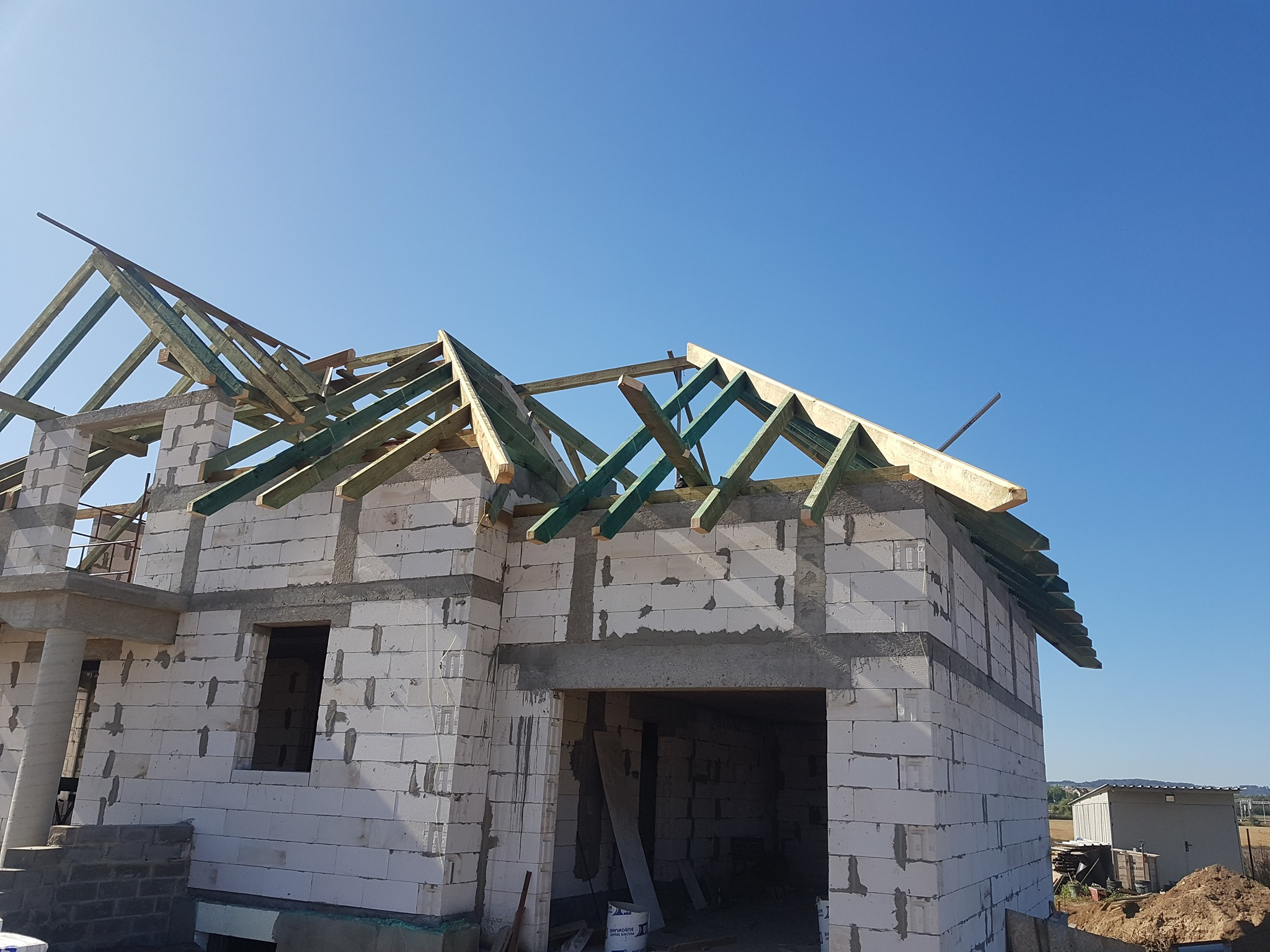 Wznoszenie domu jednorodzinnego z białych bloczków, widoczna konstrukcja drewniana dachu na tle błękitnego nieba.