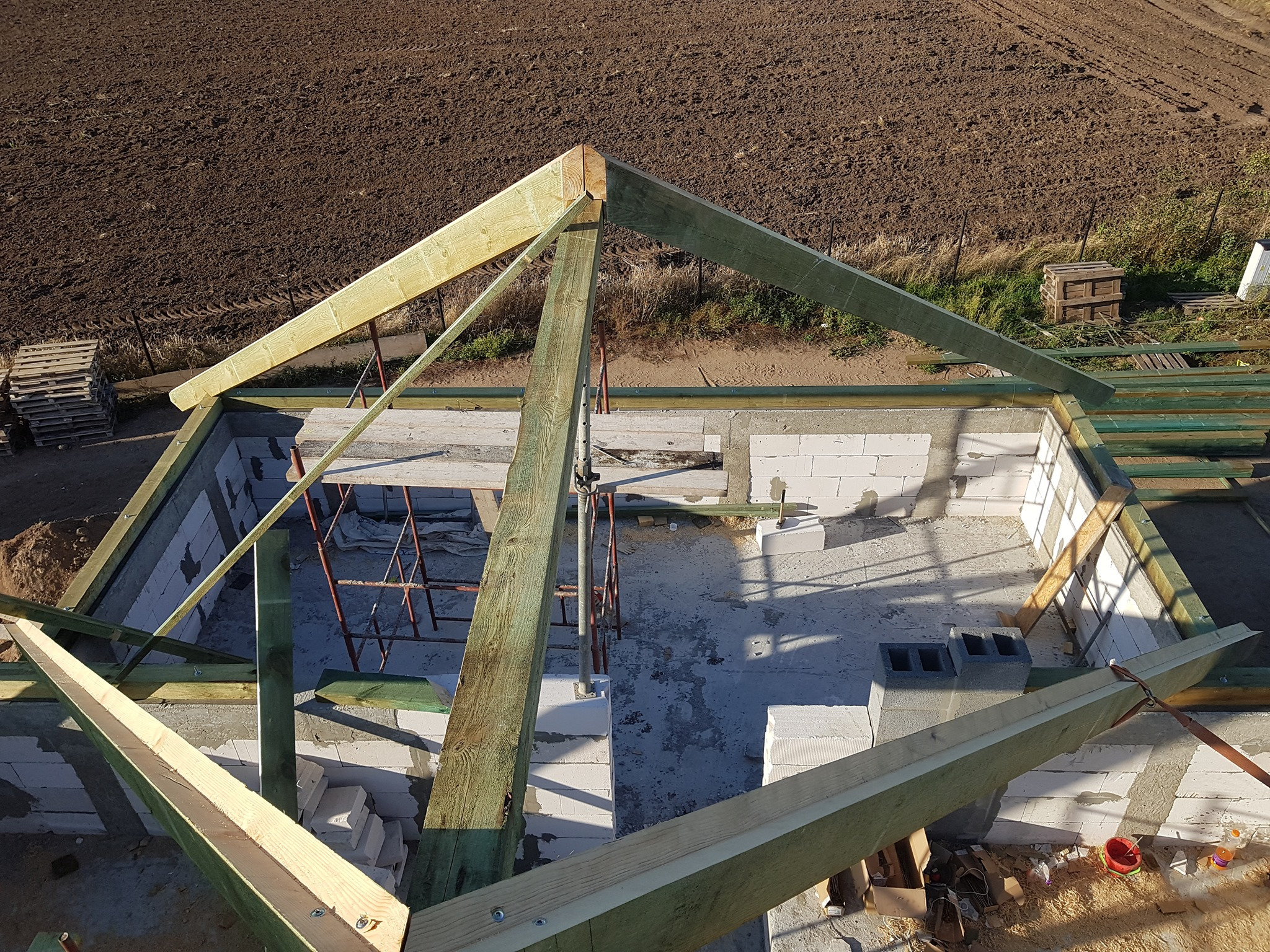 Wznoszenie drewnianej konstrukcji dachu na budynku z białych bloczków, widok z góry na niezadaszoną budowlę i pole uprawne w tle.