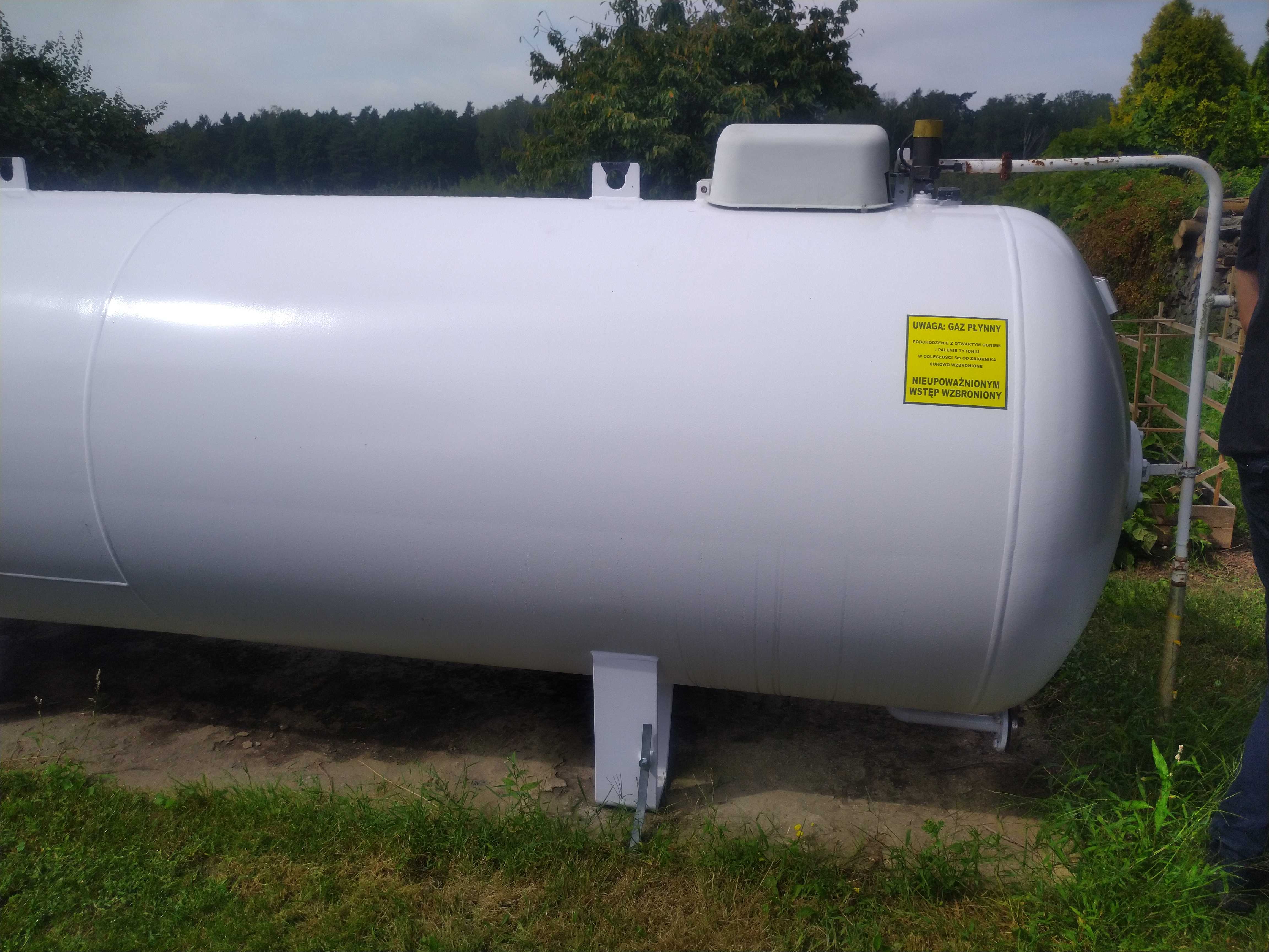 Biały, poziomy zbiornik na gaz płynny z żółtą tabliczką ostrzegawczą, umieszczony na metalowych podporach na zewnątrz, otoczony zielenią.
