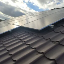 Panele słoneczne na metalowej dachówce w kolorze grafitowym, zamontowane za pomocą aluminiowych uchwytów, z widocznym fragmentem pochmurnego nieba.