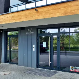 Wejście do nowoczesnego apartamentowca z szarymi drzwiami i oknami, widoczny numer budynku '47 Jesionowa' oraz logo firmy ochroniarskiej na szybie.