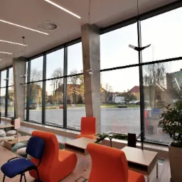 Wnętrze nowoczesnej biblioteki z dużymi oknami PCV, pomarańczowymi fotelami i widokiem na zewnątrz.