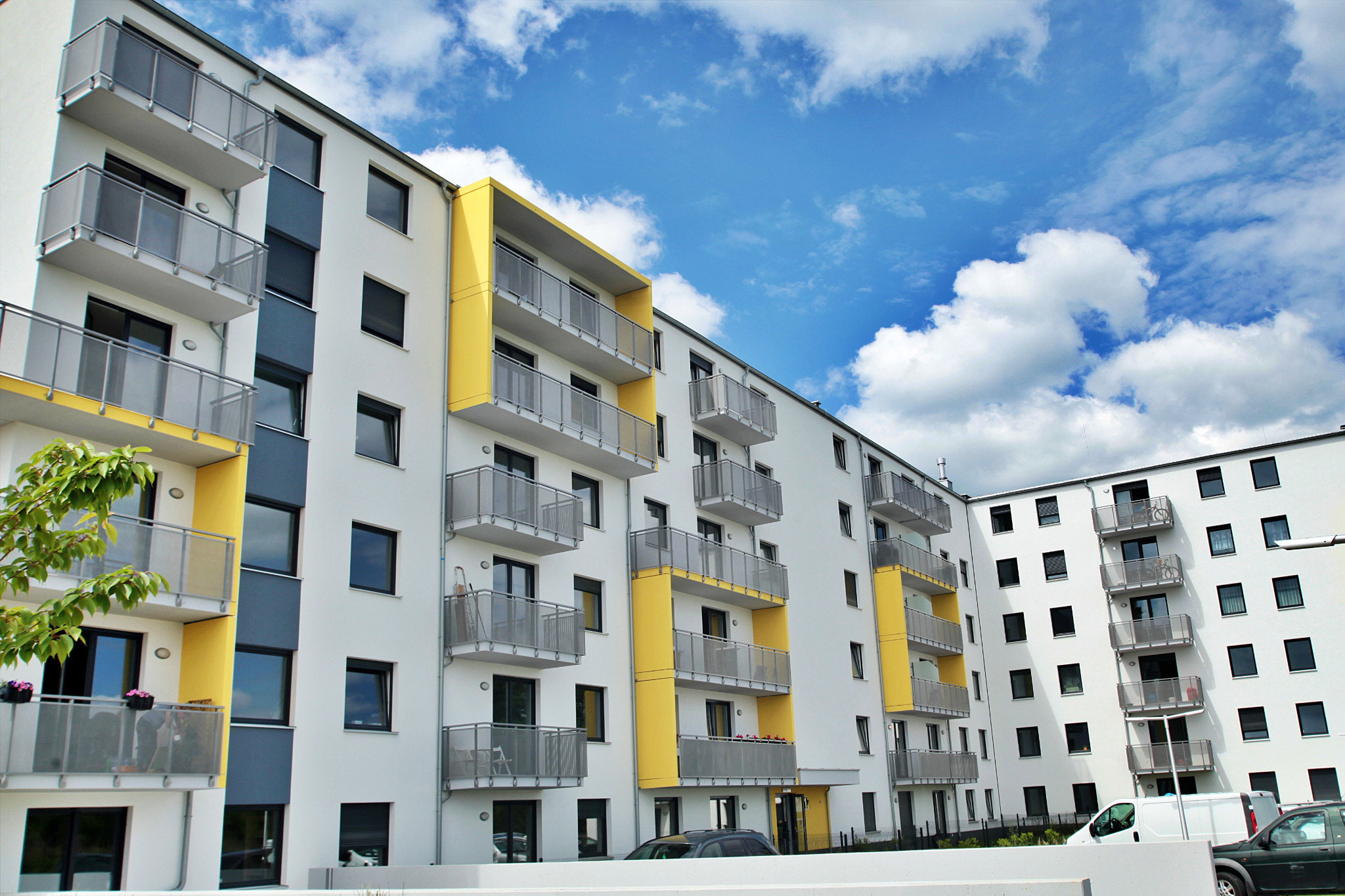 Nowoczesny budynek wielorodzinny z balkonami, białą elewacją i żółtymi akcentami architektonicznymi, na tle błękitnego nieba z białymi chmurami.
