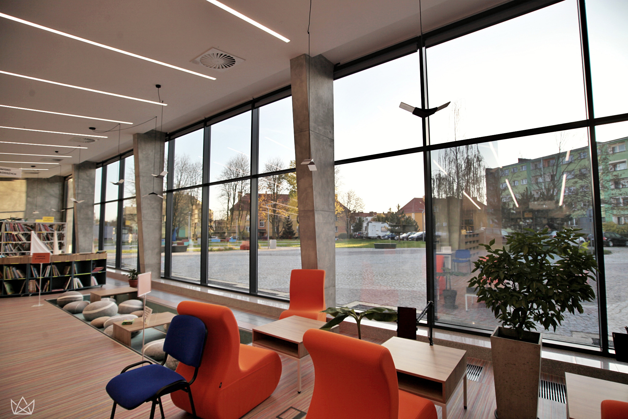 Wnętrze nowoczesnej biblioteki z dużymi oknami PCV, pomarańczowymi fotelami i widokiem na zewnątrz.