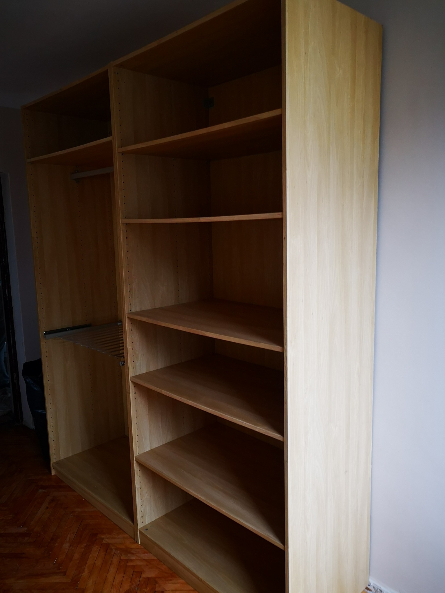 Dwa połączone regały z jasnego drewna, jeden z drążkiem i półką na ubrania, drugi z otwartymi półkami, stojące na parkiecie w pokoju z szarą ścianą.