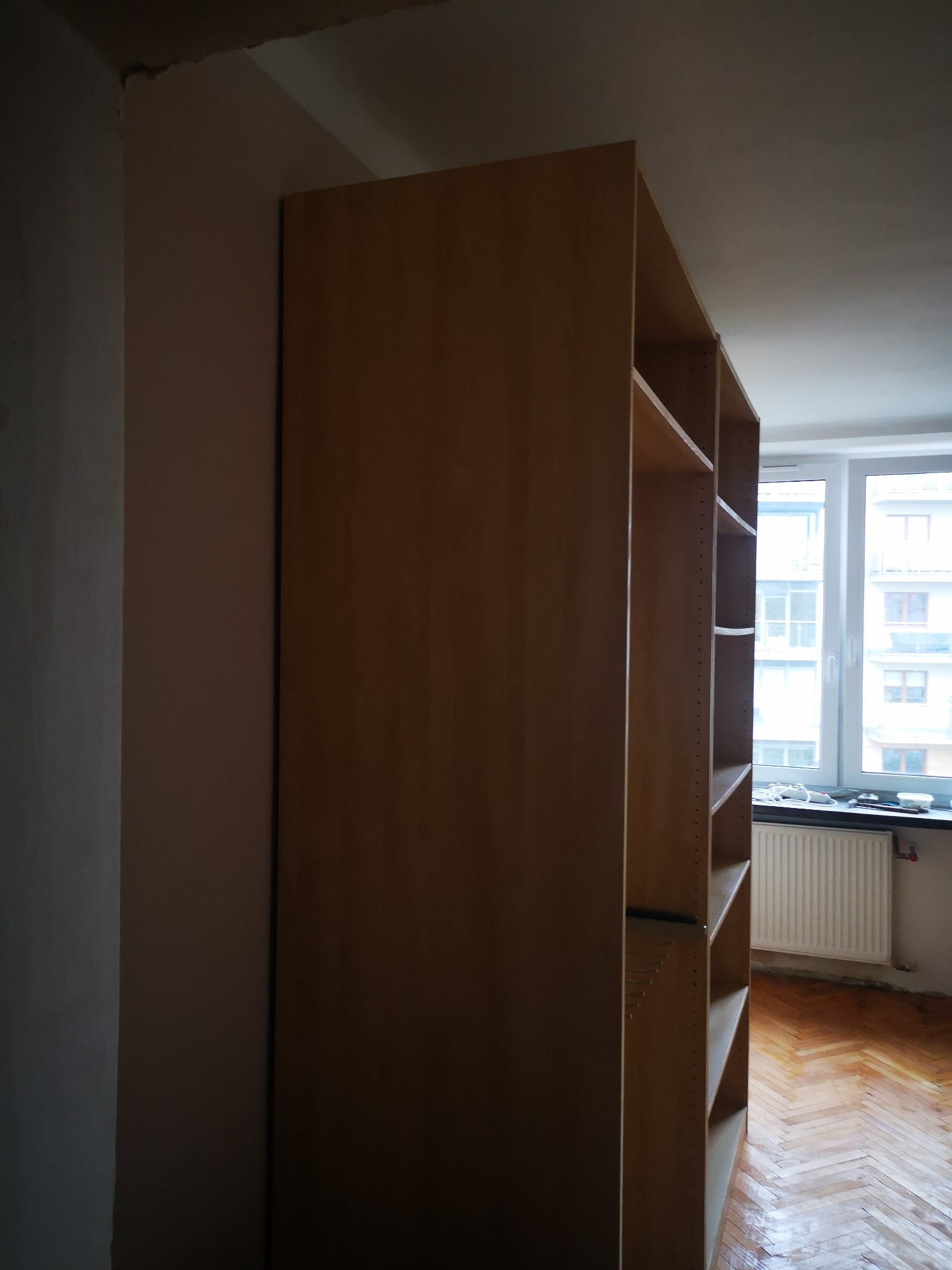 Narożny widok na wnękę z surową ścianą i częściowo zmontowany, drewniany regał na książki z widocznymi półkami, na tle okna z grzejnikiem i parkietem w jodełkę.