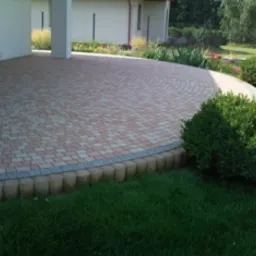 Ułożona kostka brukowa w geometryczny wzór na tarasie z obrzeżem z drewnianych palisad, widok z góry na tle budynku i zieleni.