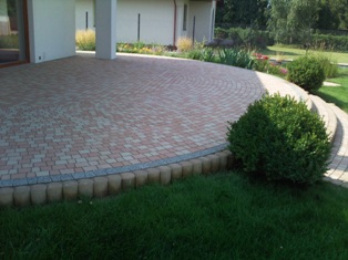 Ułożona kostka brukowa w geometryczny wzór na tarasie z obrzeżem z drewnianych palisad, widok z góry na tle budynku i zieleni.