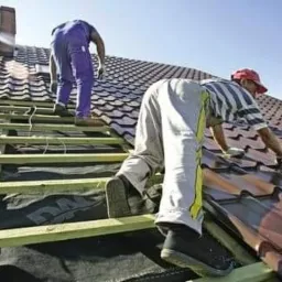 Dwóch dekarzy pracuje na dachu z brązową dachówką, jeden z nich mocuje dachówkę, drugi stoi na drewnianej konstrukcji więźby dachowej, widoczny komin w tle.