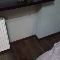 Fragment pokoju po remoncie: ciemna listwa przypodłogowa, drewnopodobne panele podłogowe, biały grzejnik i jasna ściana