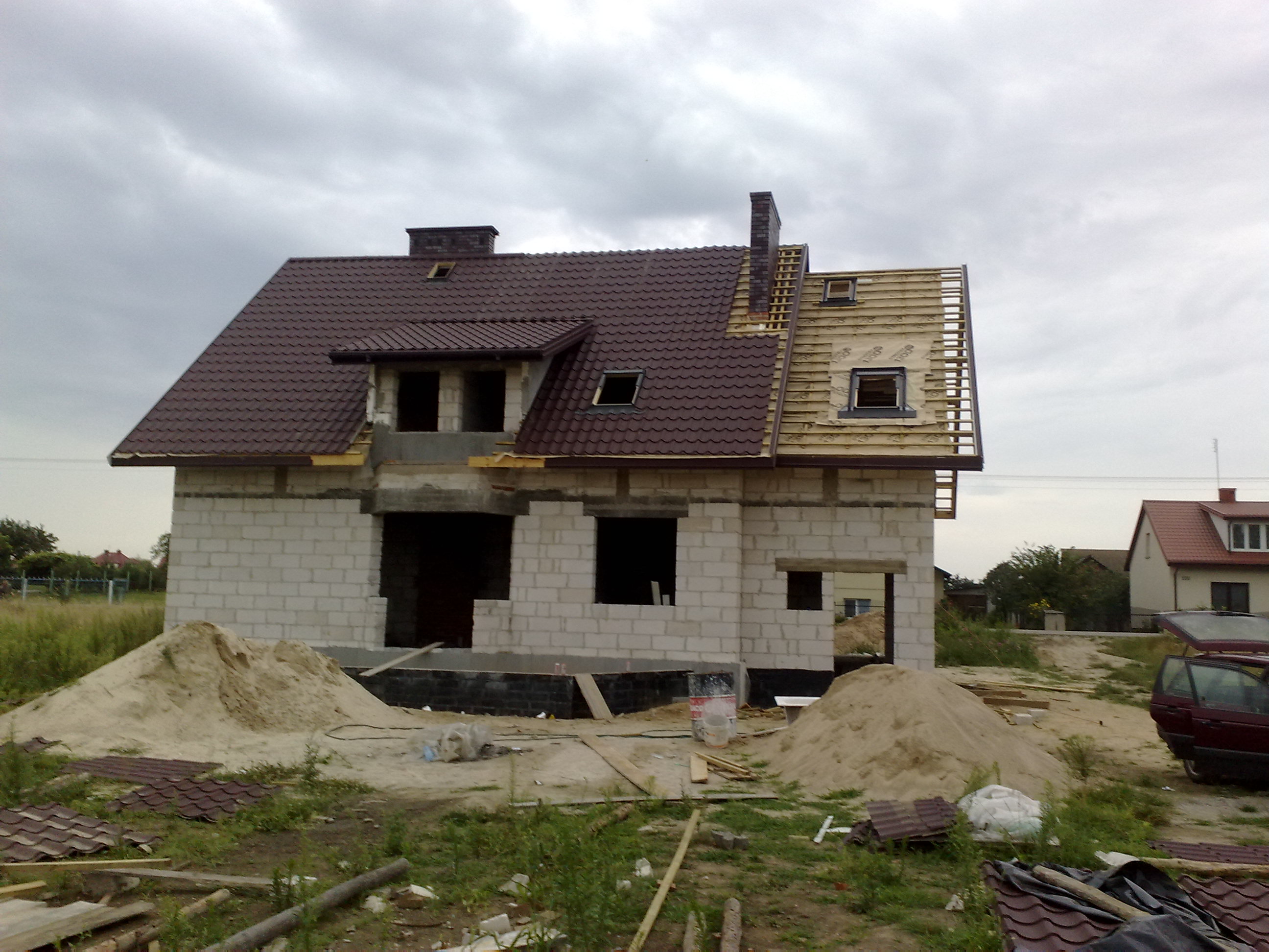 Surowy dom murowany w trakcie budowy z częściowo ukończonym dachem z brązowej blachodachówki i kominem, widoczne elementy konstrukcji drewnianej, na pierwszym planie hałdy piasku i pozostałości...