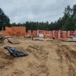 Wznoszone ściany z czerwonej cegły Porotherm na placu budowy, widoczne palety z cegłami w folii, tło stanowią drzewa i pochmurne niebo.