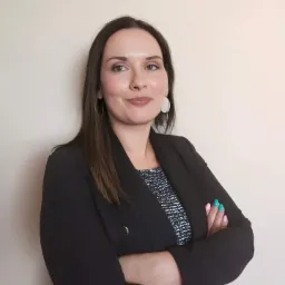 Portret kobiety w czarnej marynarce z rękami skrzyżowanymi, uśmiechniętej, z kolczykami w kształcie koła i pomalowanymi paznokciami na różowo i turkusowo.