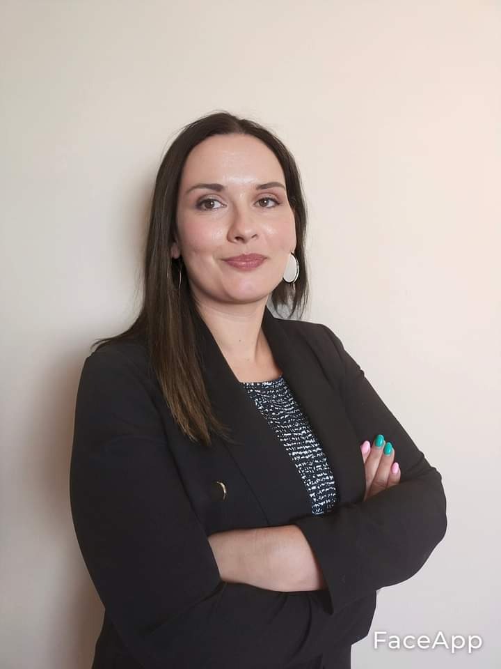 Portret kobiety w czarnej marynarce z rękami skrzyżowanymi, uśmiechniętej, z kolczykami w kształcie koła i pomalowanymi paznokciami na różowo i turkusowo.