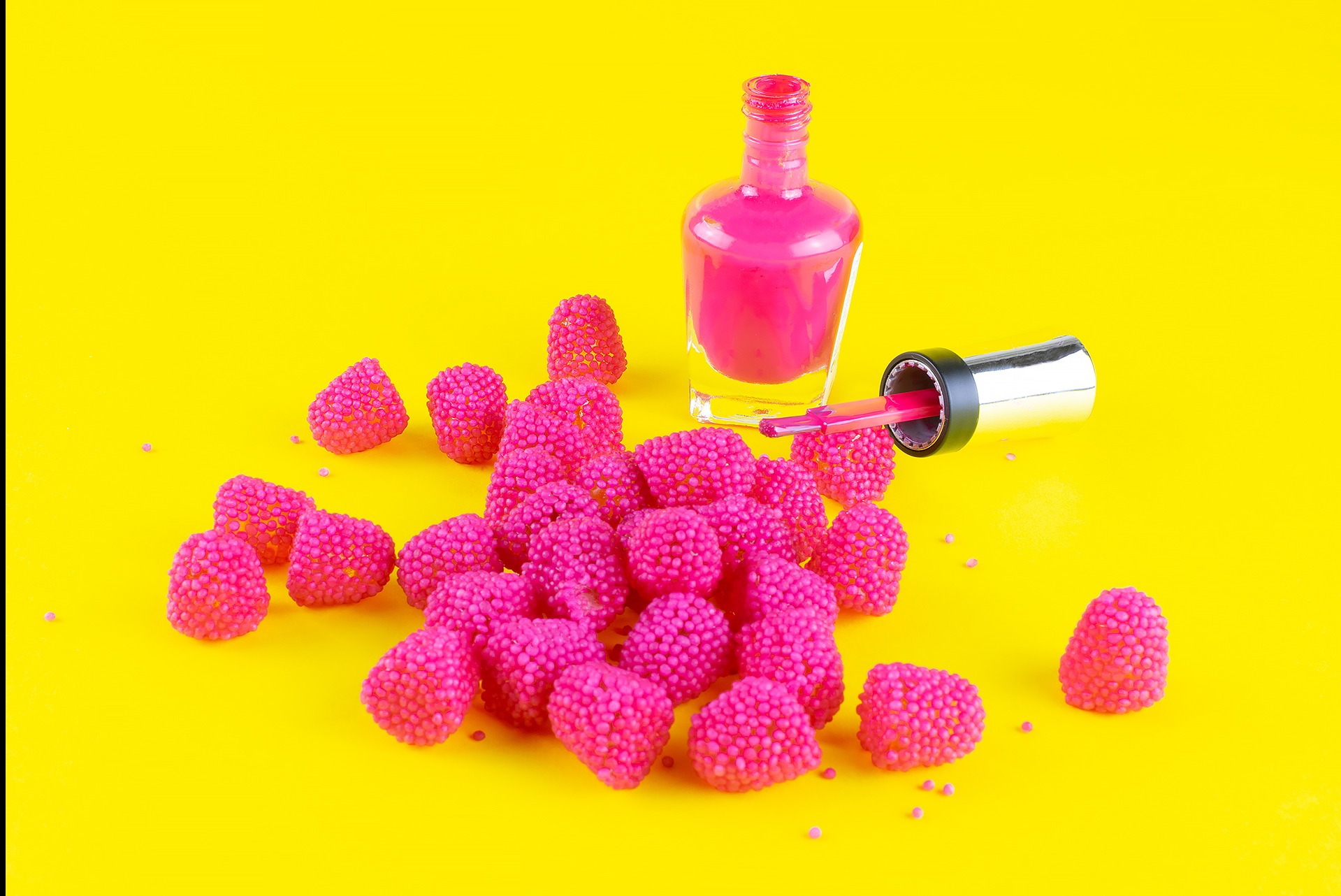 Butelka różowego lakieru do paznokci z pędzelkiem i otoczona żelkami w kształcie malin na jaskrawożółtym tle w Warszawie.