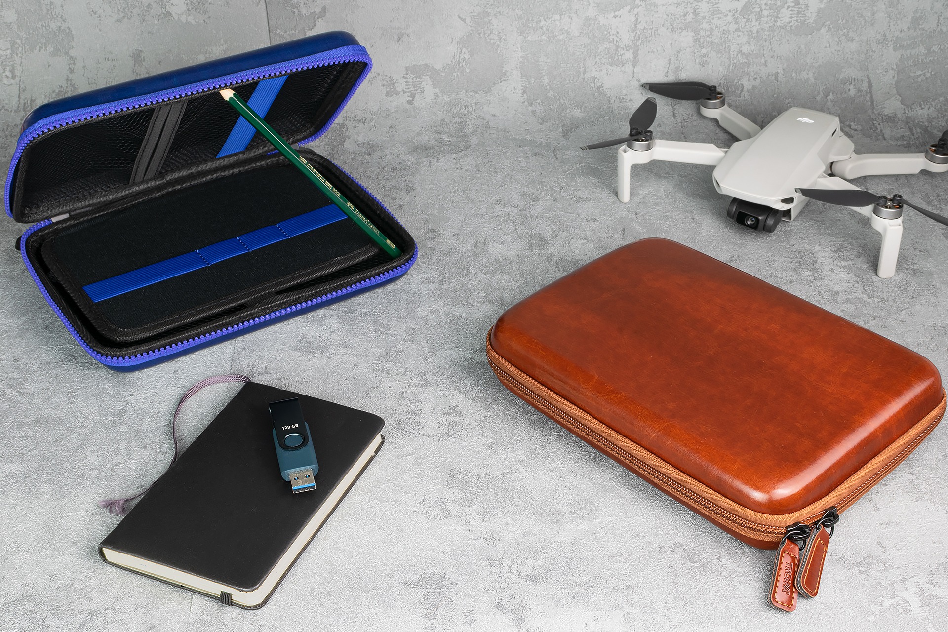 Dwa futerały zapinane na zamek, biały dron, pendrive na notesie i ołówek w niebieskim etui na szarym tle.