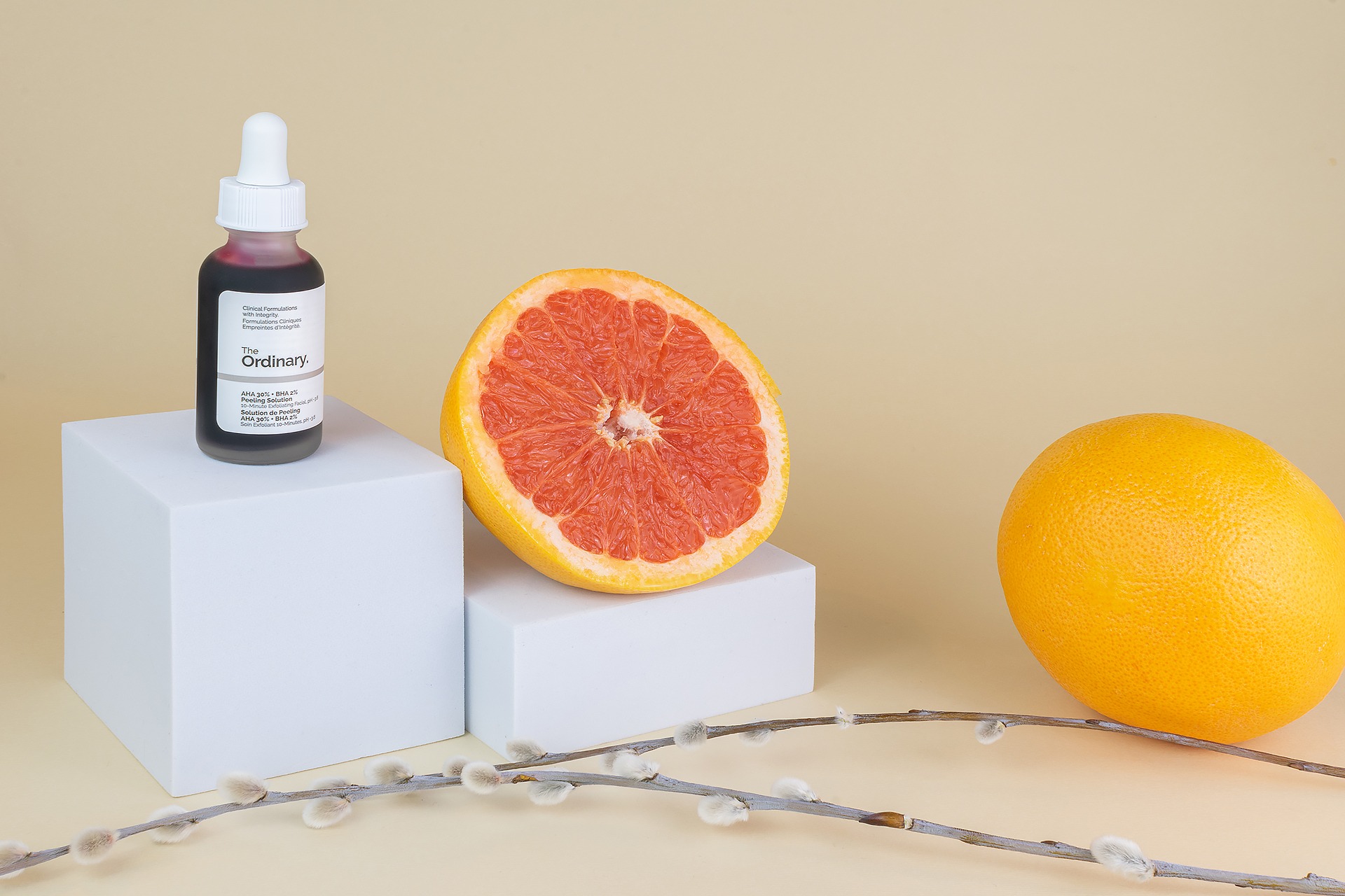 Butelka serum do twarzy The Ordinary obok przekrojonego grejpfruta i pomarańczy na białych podestach z gałązką wierzby z baziami na beżowym tle.