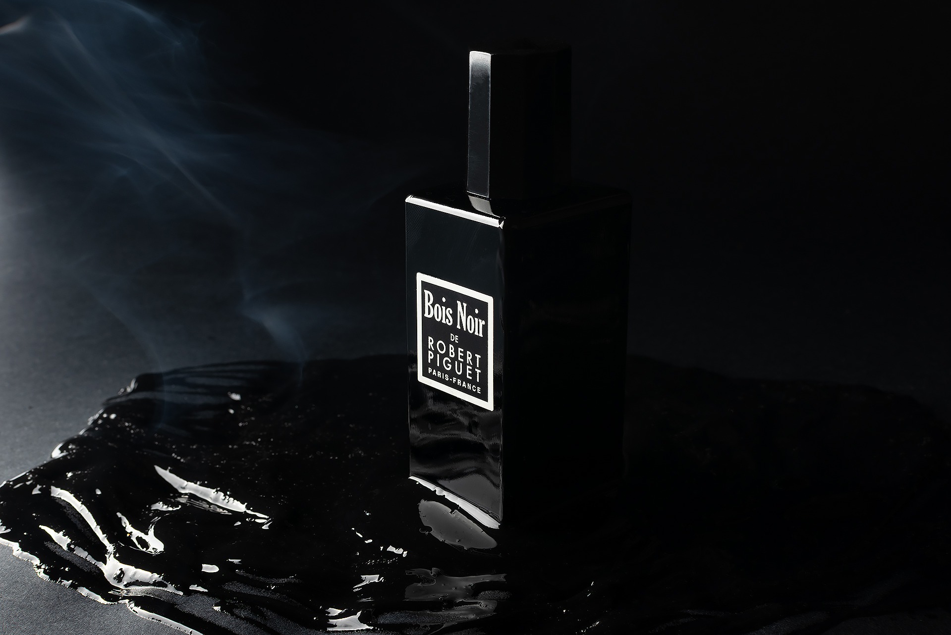Czarny flakon perfum Bois Noir de Robert Piguet otoczony smugami dymu i czarną, błyszczącą substancją na ciemnym tle.