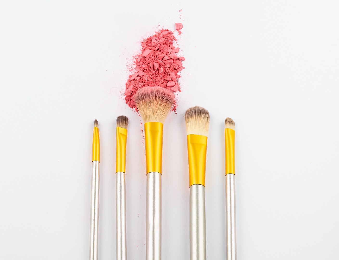 Róż do policzków w odcieniach koralowych rozsypany obok zestawu pędzli do makijażu z żółtymi uchwytami na białym tle.