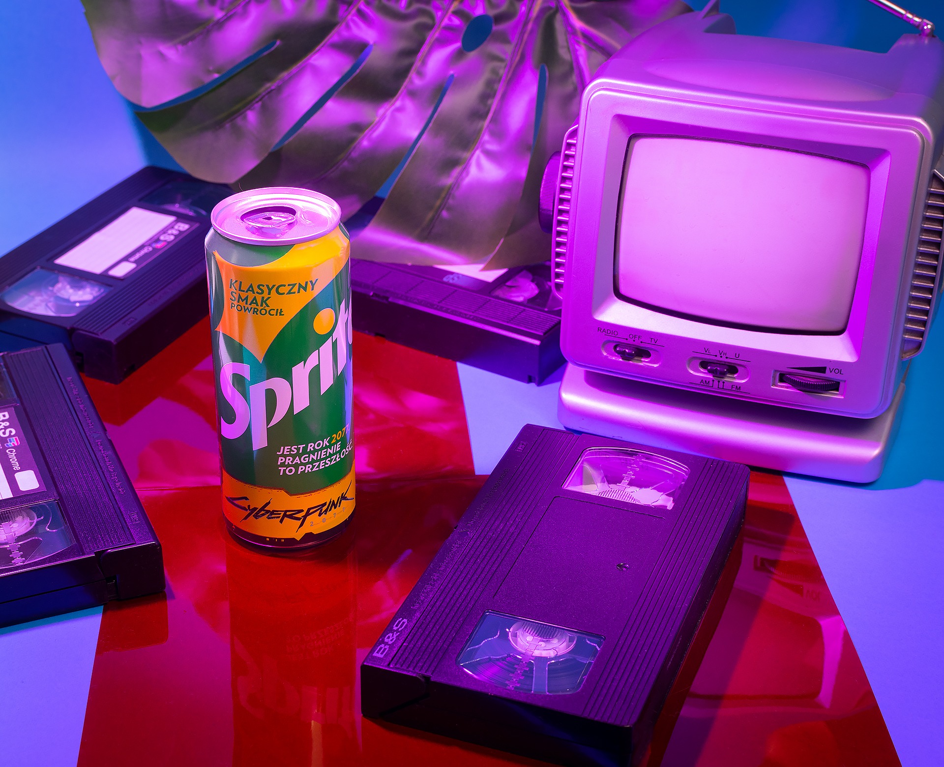 Retro still life: puszka Sprite Cyberpunk 2077, kasety VHS i srebrny telewizor CRT podświetlone neonowym fioletem i błękitem, z liściem monstery w tle.