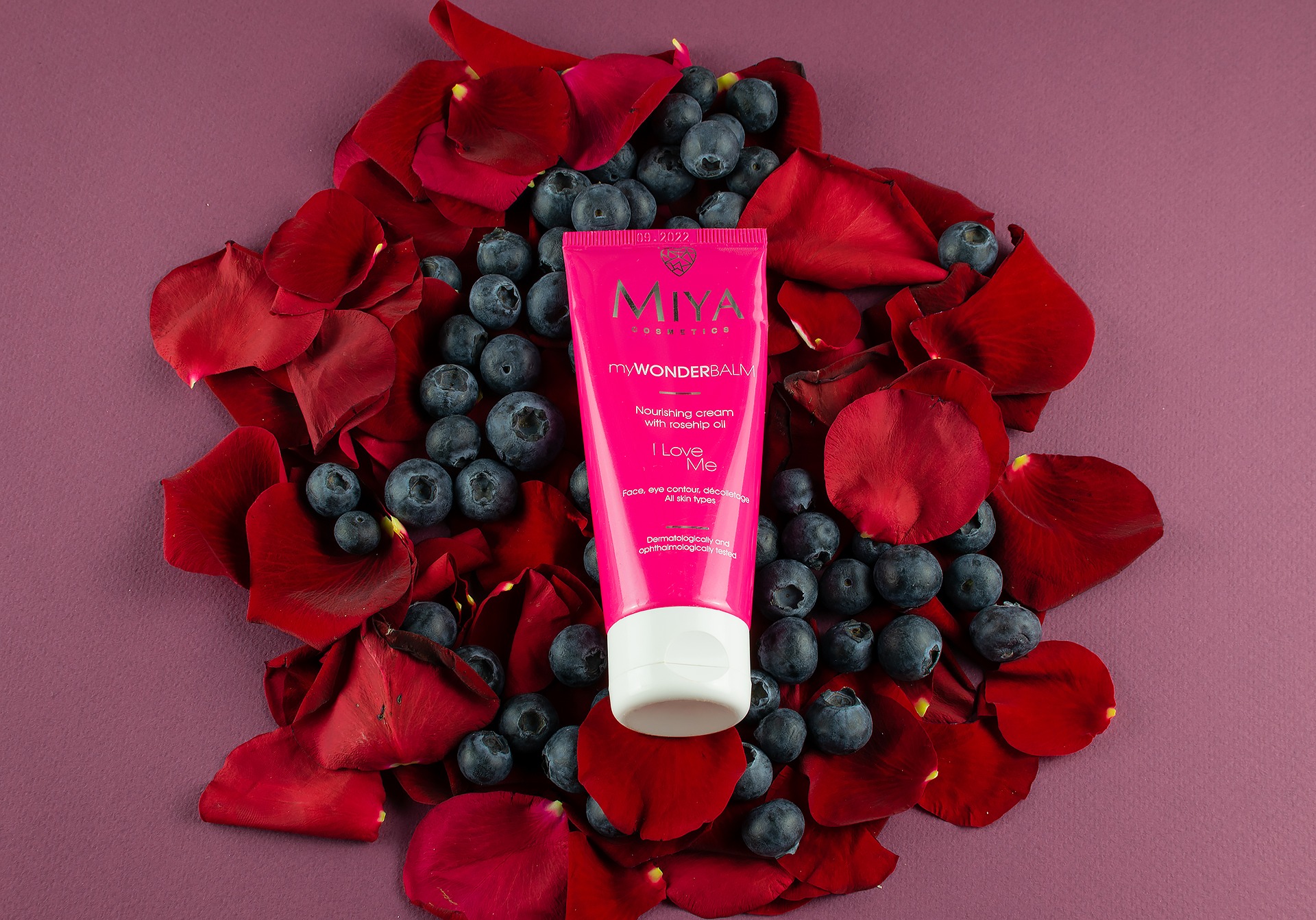 Różowa tuba kremu Miya myWONDERBALM na tle płatków czerwonej róży i borówek, ułożona na fioletowym tle.