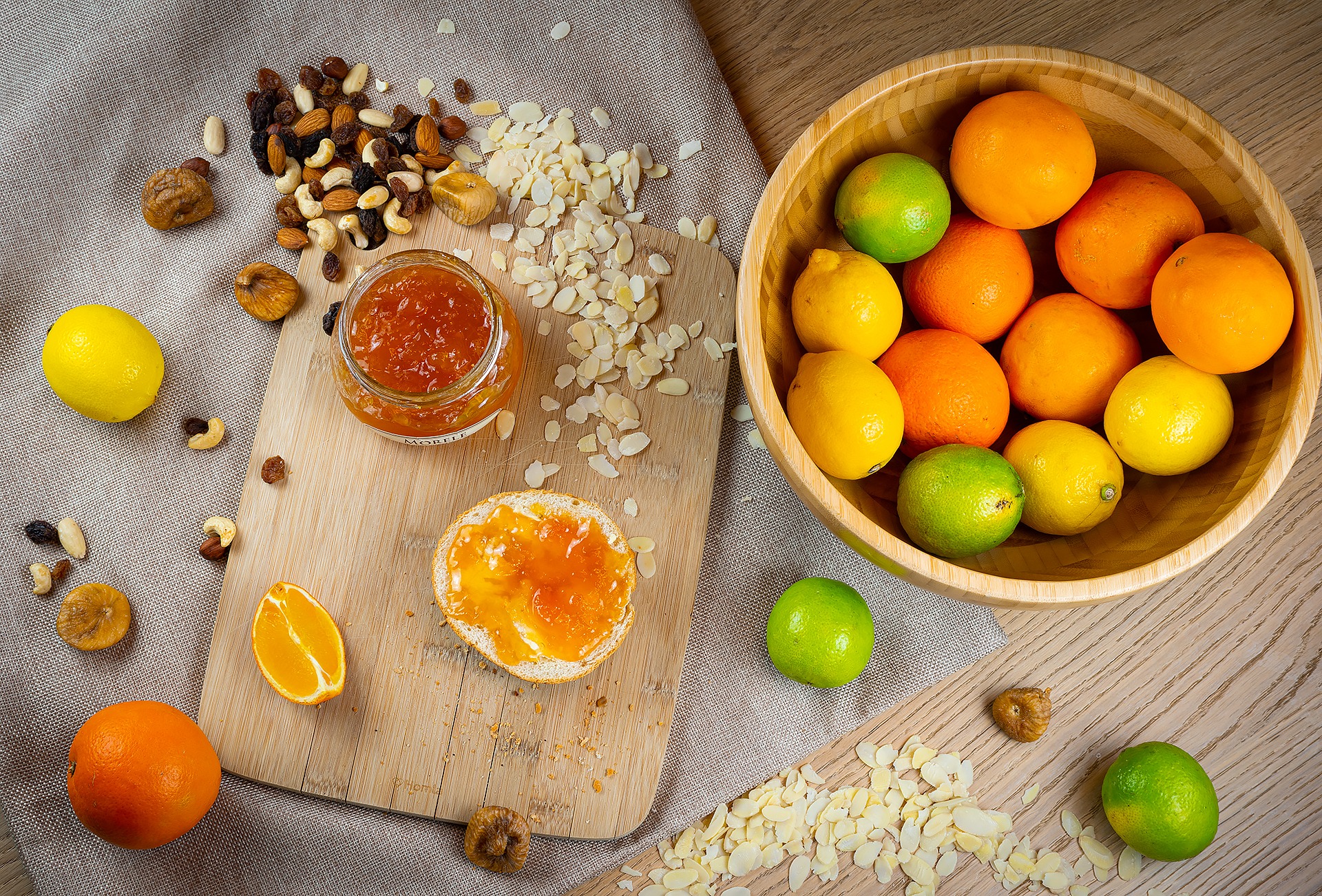 Kompozycja z cytrusów w drewnianej misce, słoika dżemu morelowego, kromki chleba z dżemem, orzechów, fig i płatków migdałów na drewnianej desce i szarej ściereczce.