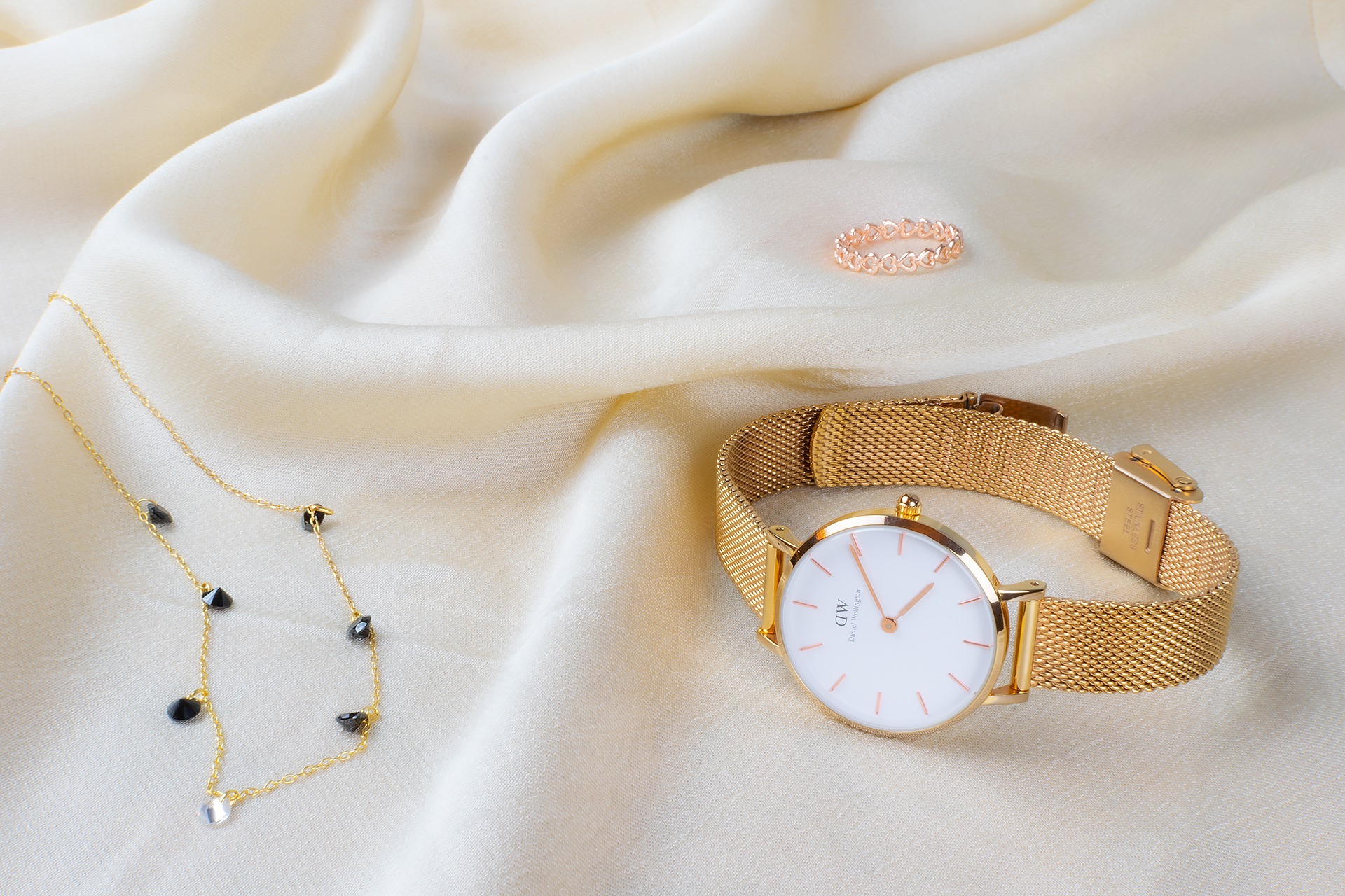 Złoty zegarek na bransolecie typu mesh, pierścionek z motywem serc i złoty naszyjnik z czarnymi kamieniami na kremowym, jedwabnym tle.