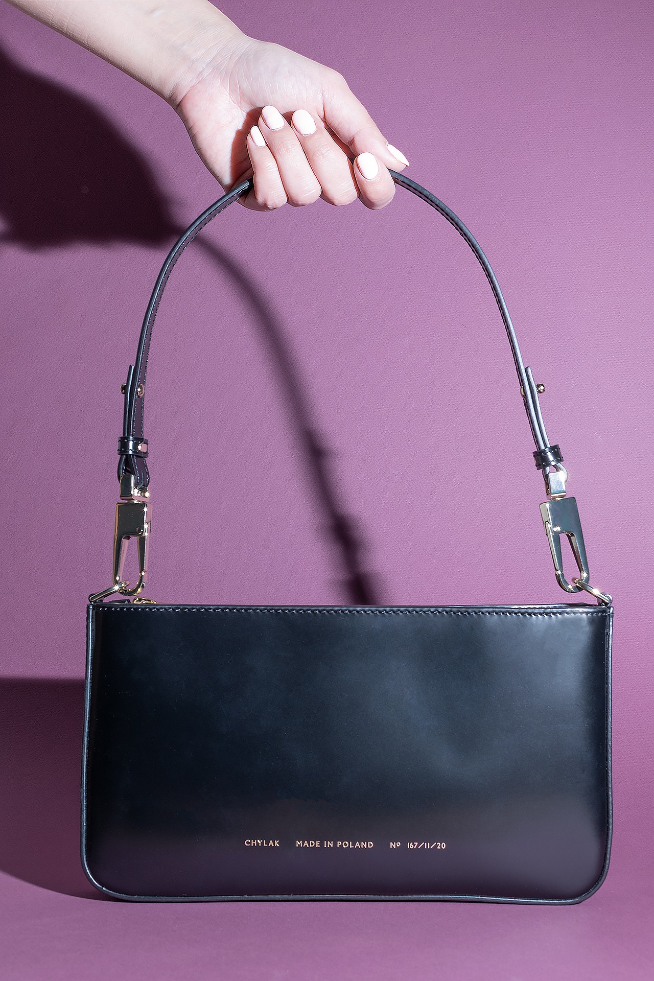 Czarna, lakierowana torebka marki CHYLAK, z napisem 'MADE IN POLAND' i numerem seryjnym, trzymana w dłoni na tle fioletowej ściany.