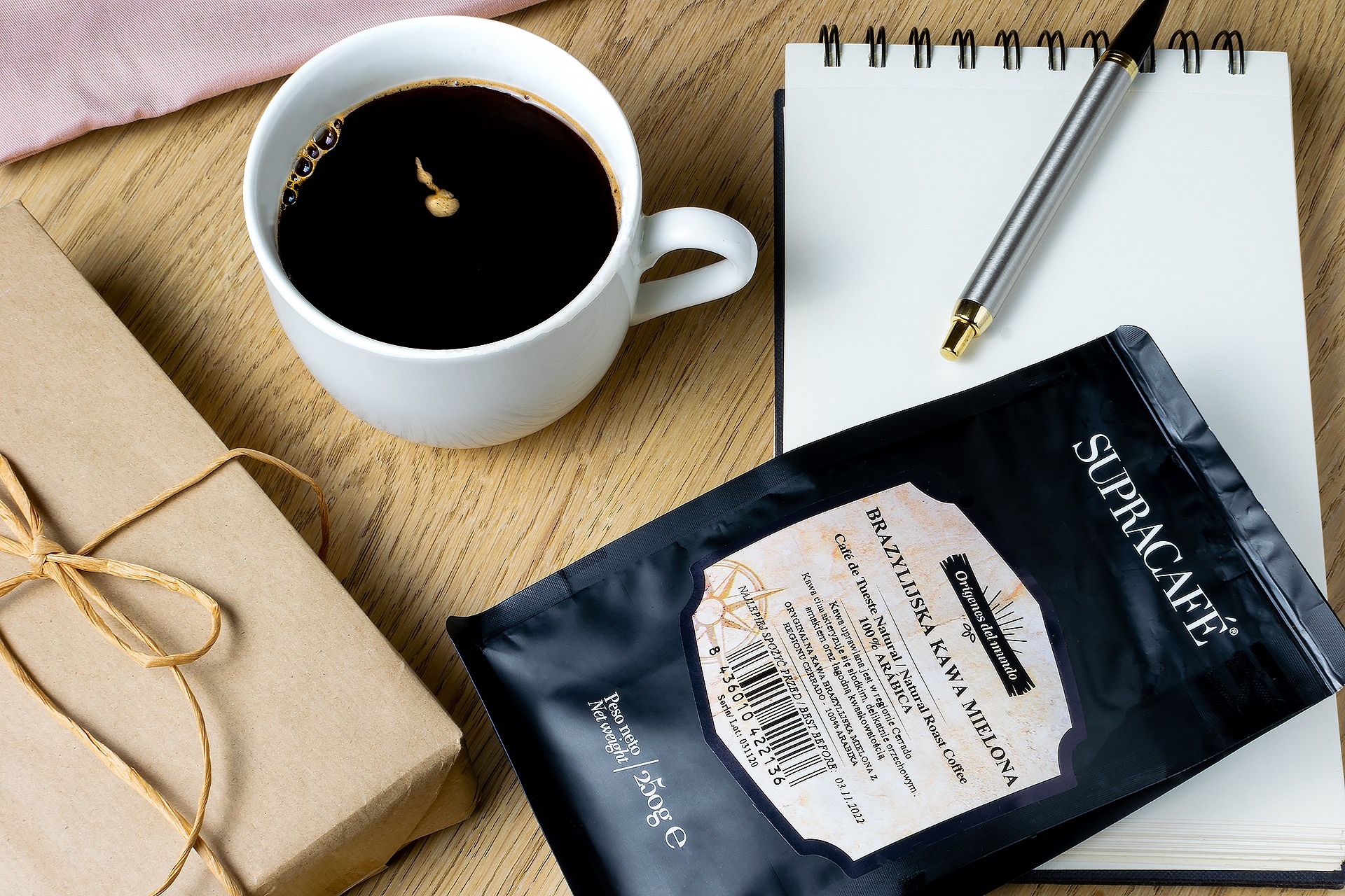Filiżanka czarnej kawy, paczka kawy brazylijskiej, notatnik z długopisem i prezent zapakowany w brązowy papier na drewnianym stole.