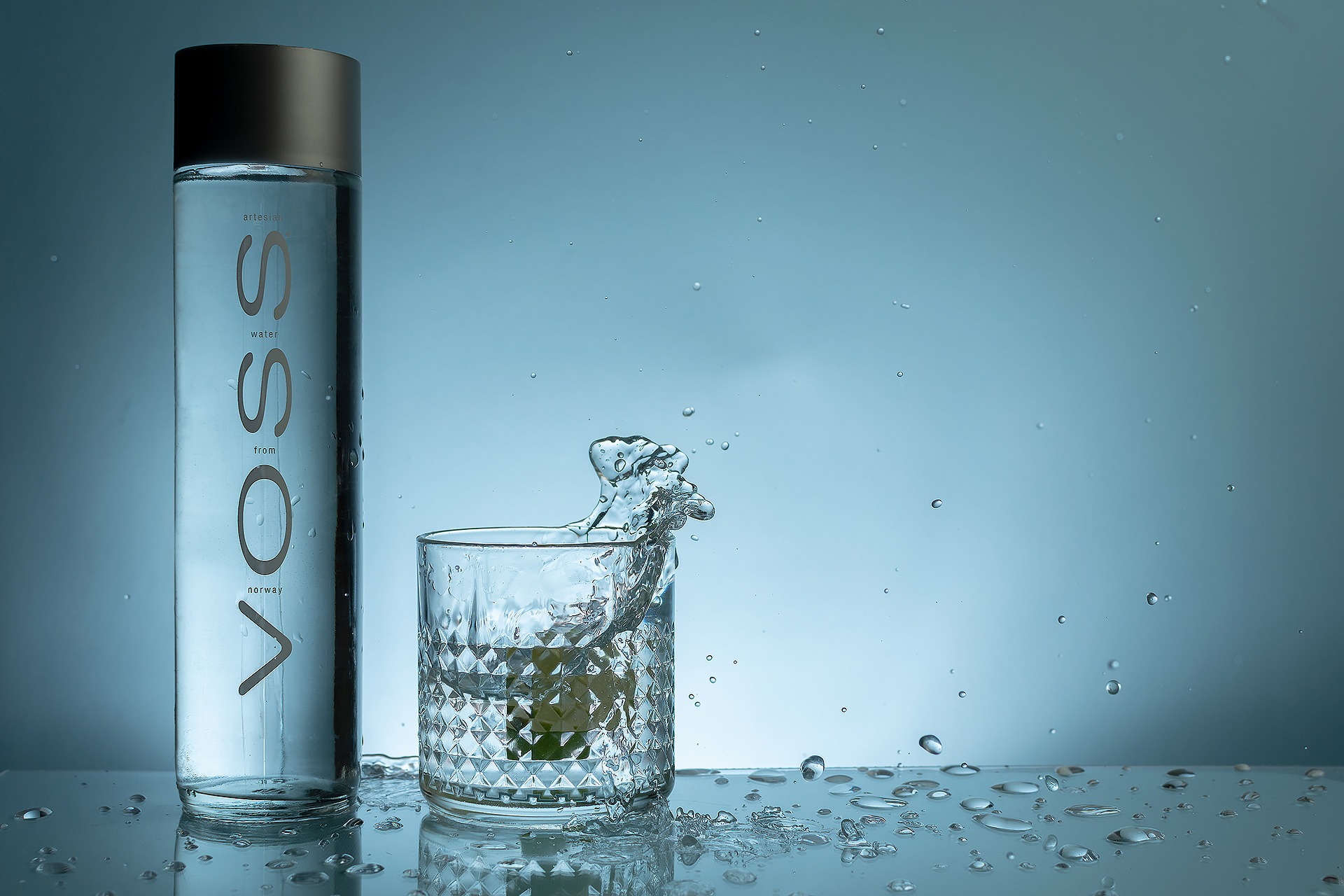 Butelka wody Voss obok szklanki z kostką lodu, w której woda rozpryskuje się, tworząc dynamiczną scenę na jasnoniebieskim tle z kroplami wody.