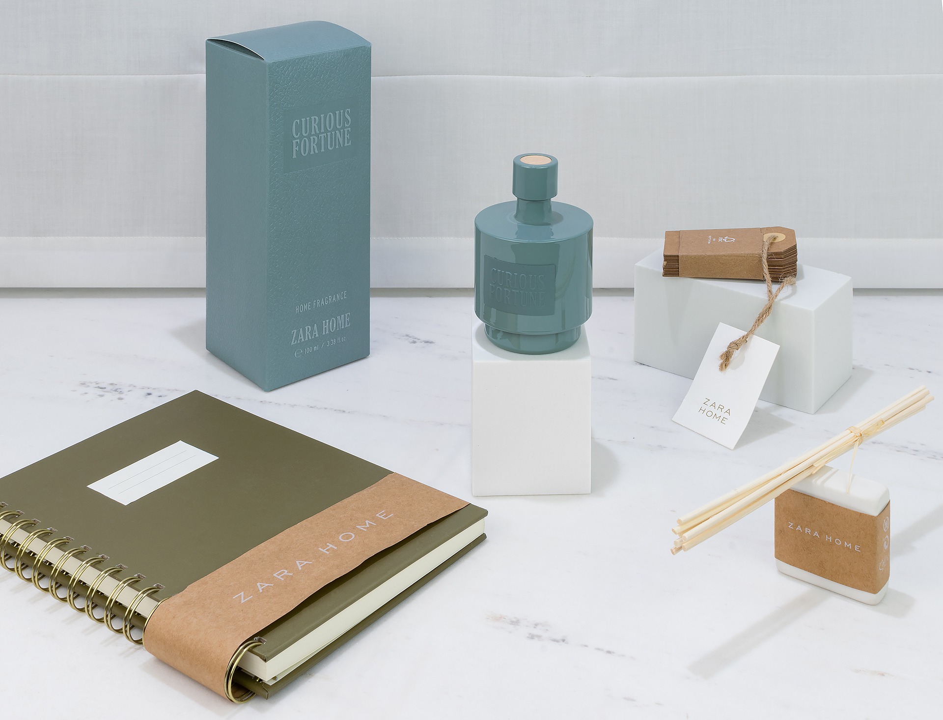 Kompozycja produktowa Zara Home: turkusowy flakon perfum Curious Fortune na białym postumencie, pudełko, notatnik w kolorze khaki z banderolą, dyfuzor zapachowy z patyczkami, wszystko na marmurowym...
