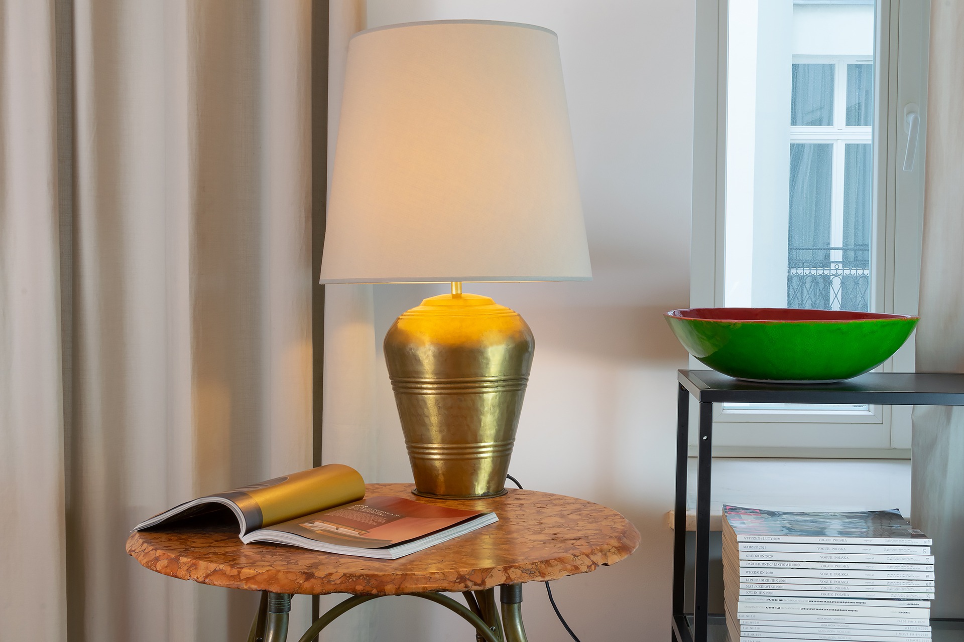 Lampa z mosiężną podstawą i beżowym abażurem na okrągłym stoliku z marmurowym blatem, obok otwarta gazeta, w tle okno i zielona misa na czarnym regale.