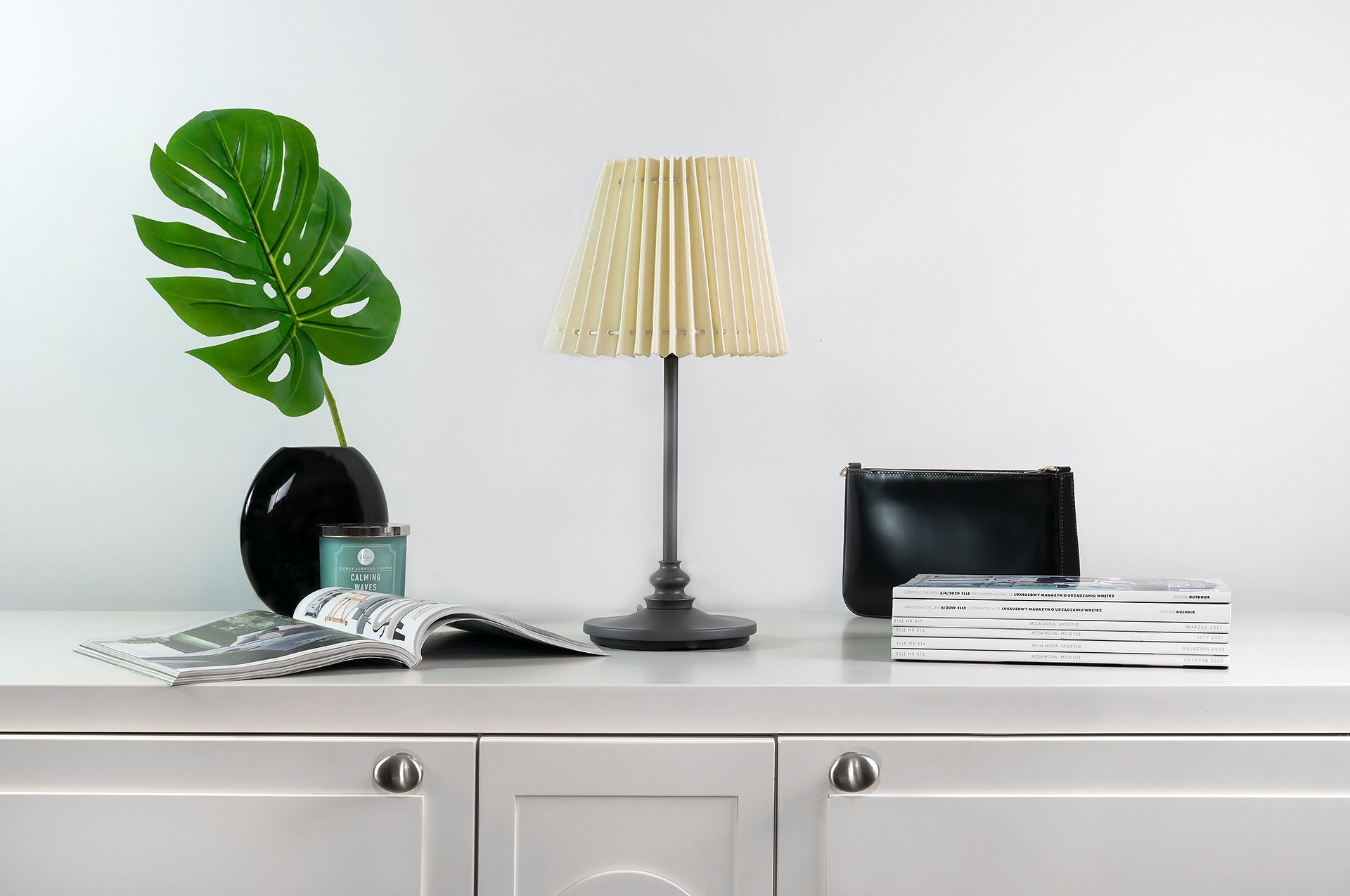 Aranżacja wnętrza: czarny wazon z liściem monstery, świeca zapachowa, rozłożony magazyn, lampka nocna z plisowanym abażurem i czarna kosmetyczka obok stosu magazynów na białej komodzie z szufladami.
